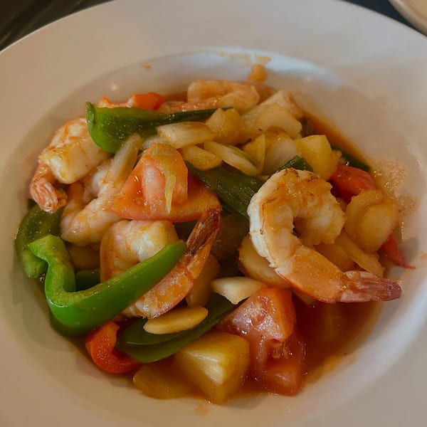 Sweet and sour shrimp with pineapple, bell peppers, tomatoes, and onions in a white bowl.