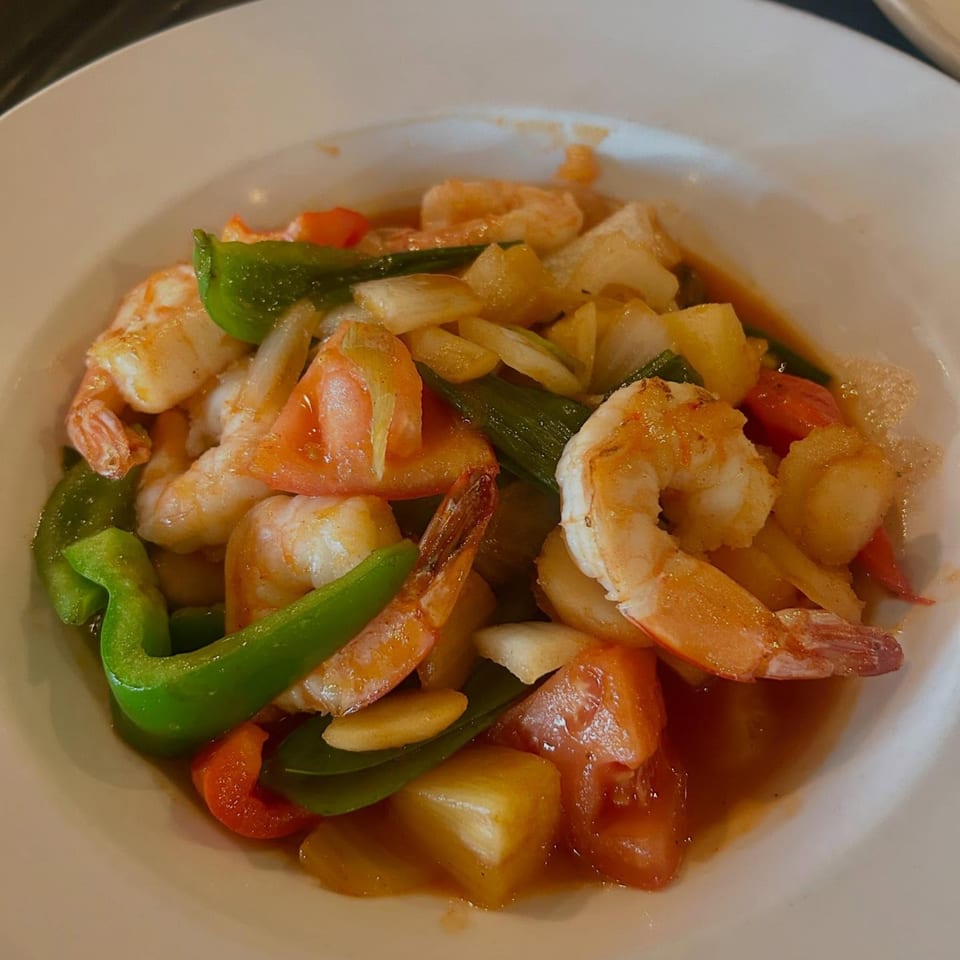 Sweet and sour shrimp with pineapple, bell peppers, tomatoes, and onions in a white bowl.