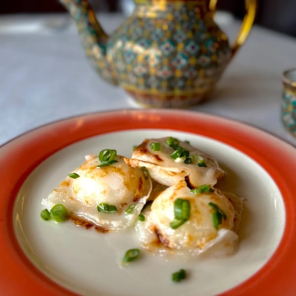 Three Thai dumplings topped with green onions on a white and red plate, with a patterned teapot in the background.