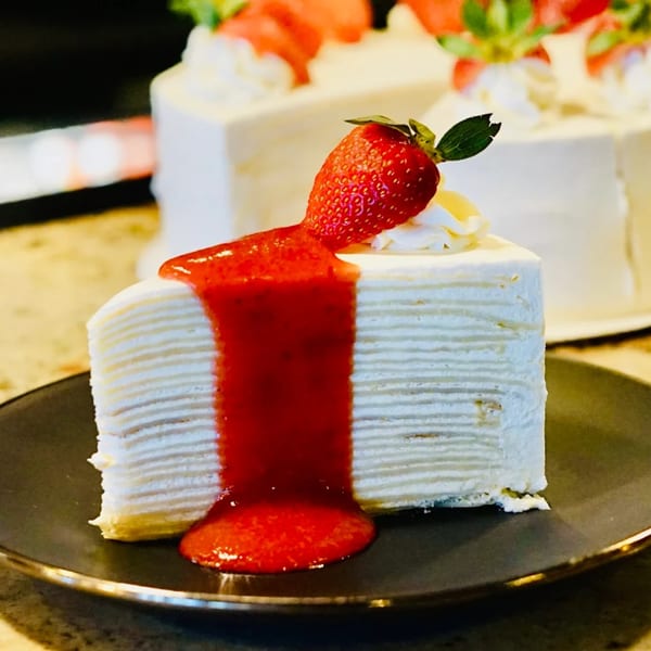 A slice of crepe cake with strawberry topping and a whole strawberry on a dark plate.